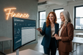 Two women in business attire standing next to a large screen displaying financial data in an office setting.